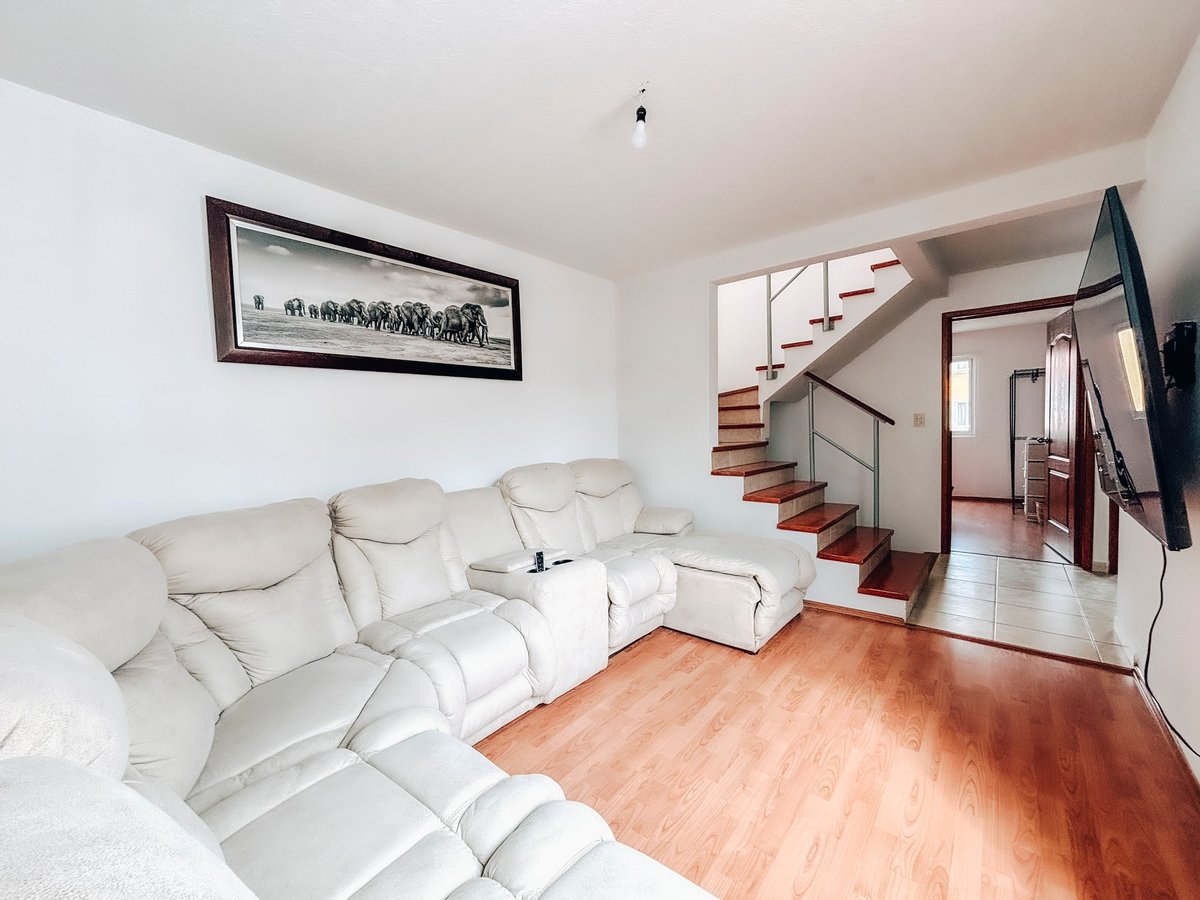 Modern living room with white sectional sofa, wooden floor, framed artwork, TV mounted on wall, and staircase leading to upper level
