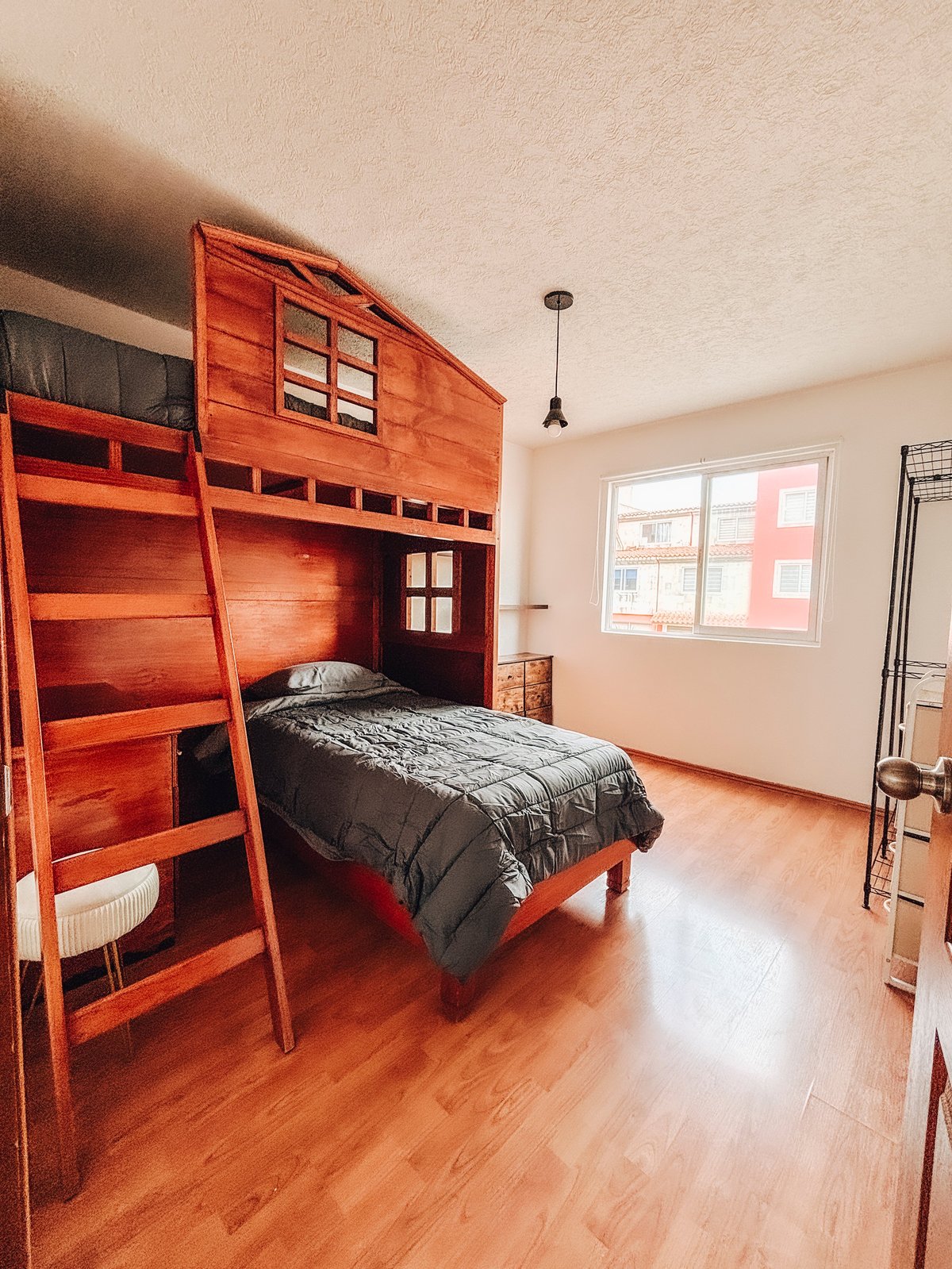 Bedroom with wooden loft bed featuring black bedding, ladder access, and desk space, large window overlooking residential building