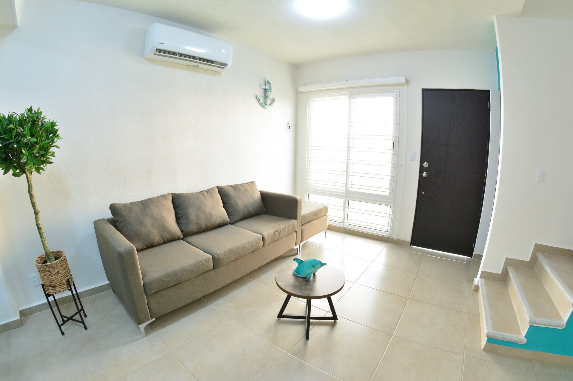 Modern living room with beige sofa, glass side table, green plant, black door, and bright white walls with skylights