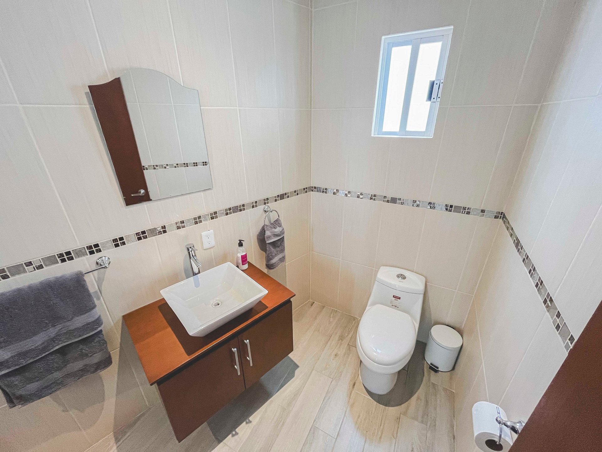 Modern bathroom interior with white tile walls, wooden vanity with sink, toilet, small window, and decorative tile border