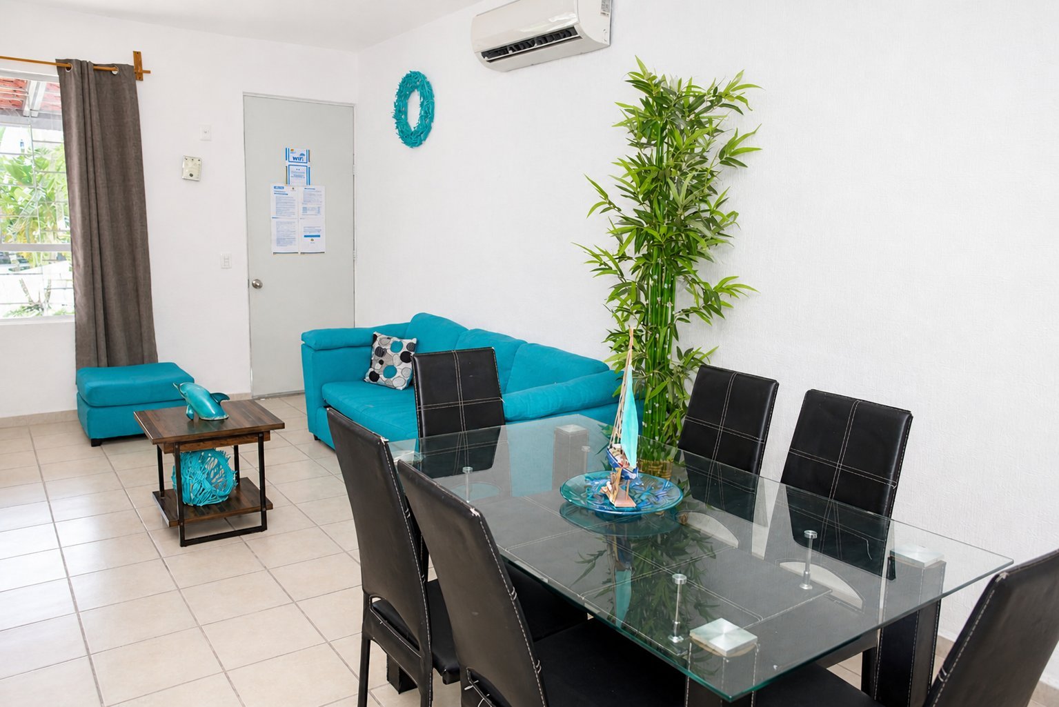 Modern living room with turquoise sectional sofa, black dining table with chairs, potted bamboo plant, and large window with curtains.