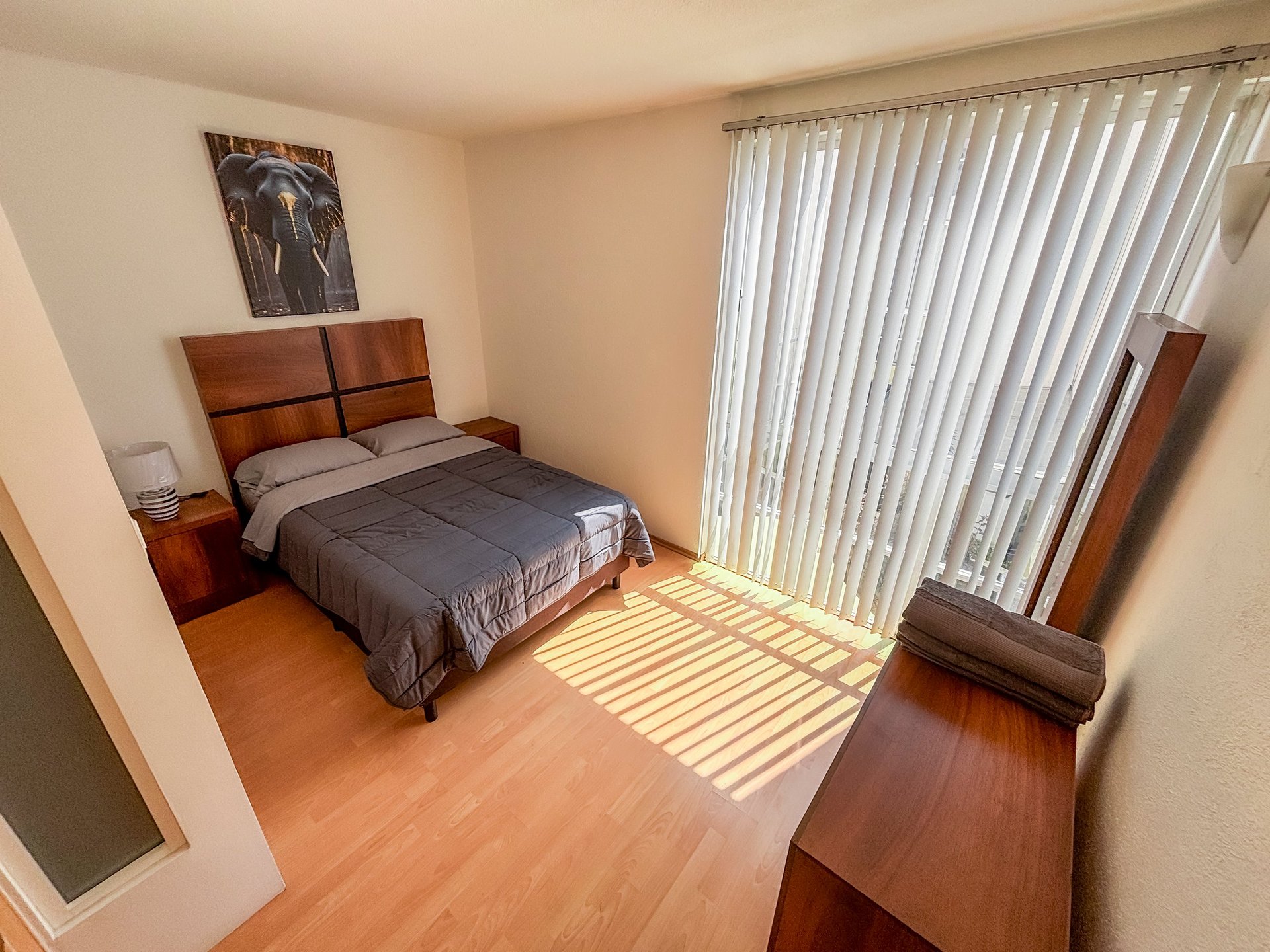 Modern bedroom with wooden bed frame, dark bedding, hardwood floors, and vertical blinds on large window