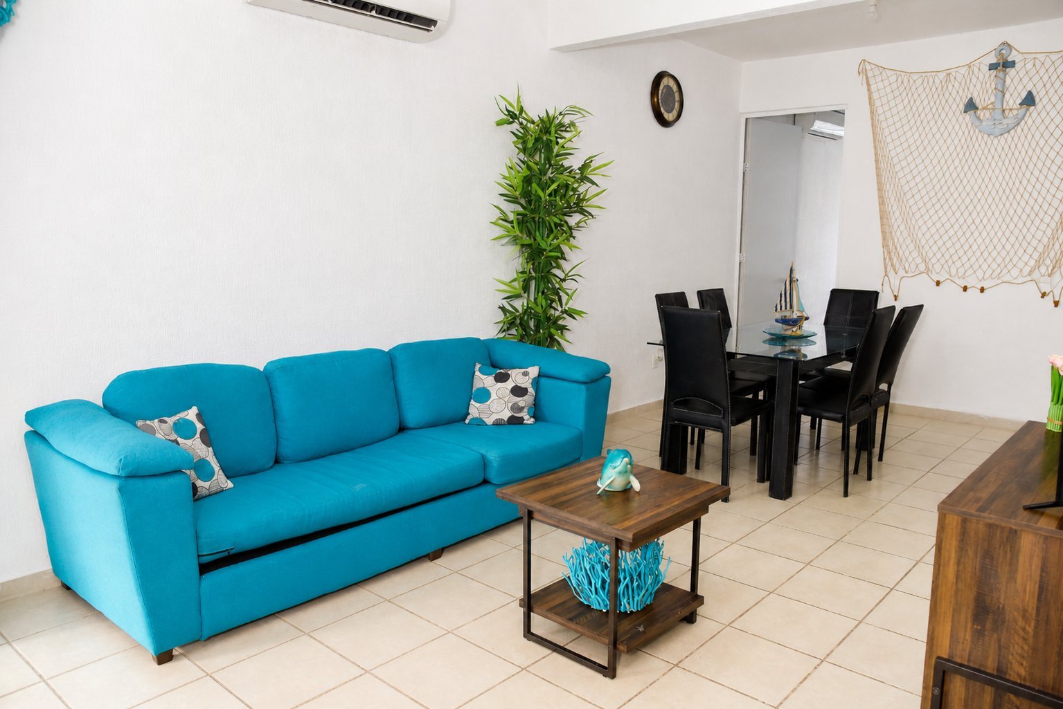 Modern living room with bright blue sofa, wooden coffee table, black dining set, and green plant accents.