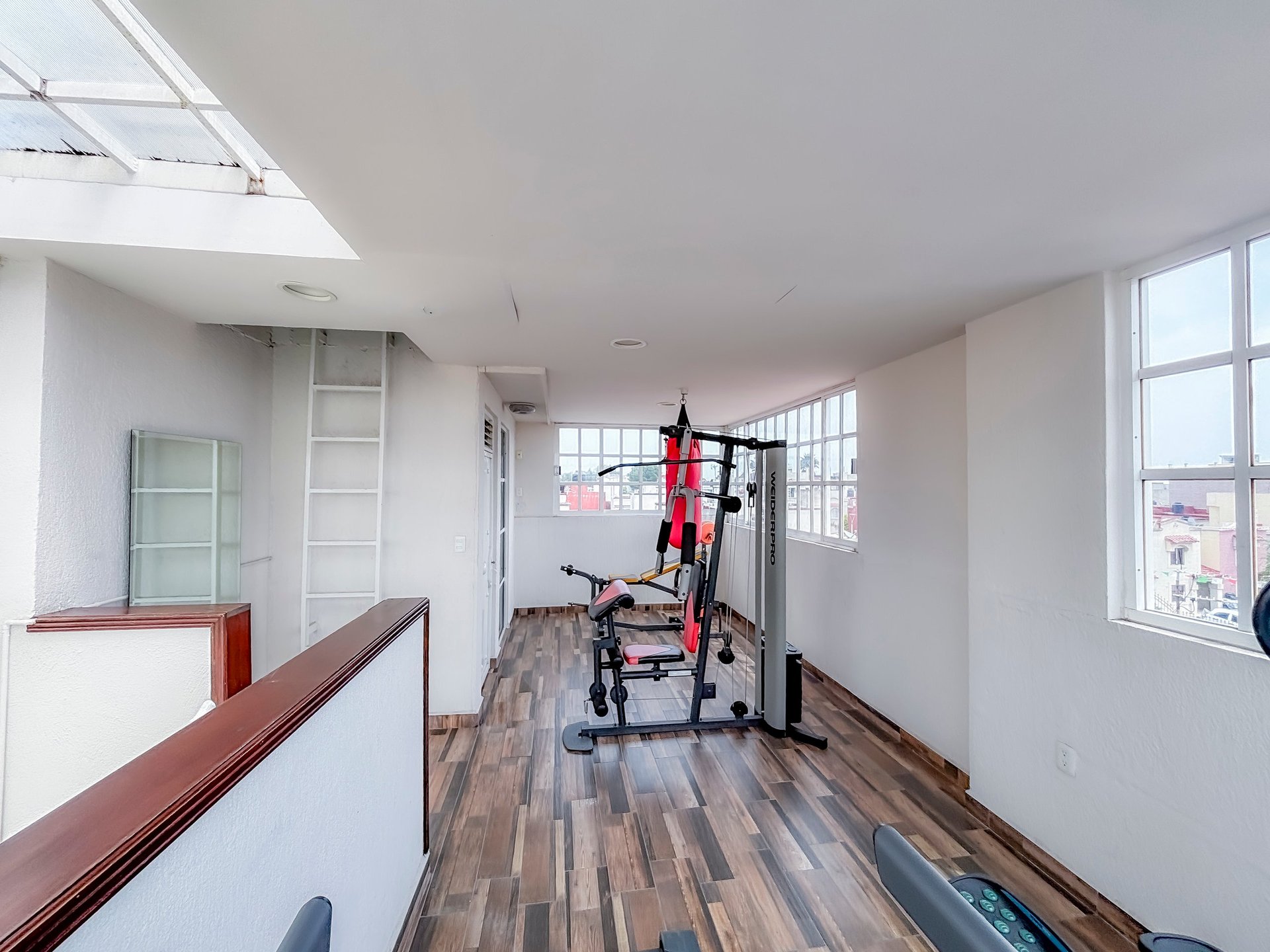 Modern loft interior with red stationary bike, wooden floors, skylights, and white walls with large windows overlooking a city.