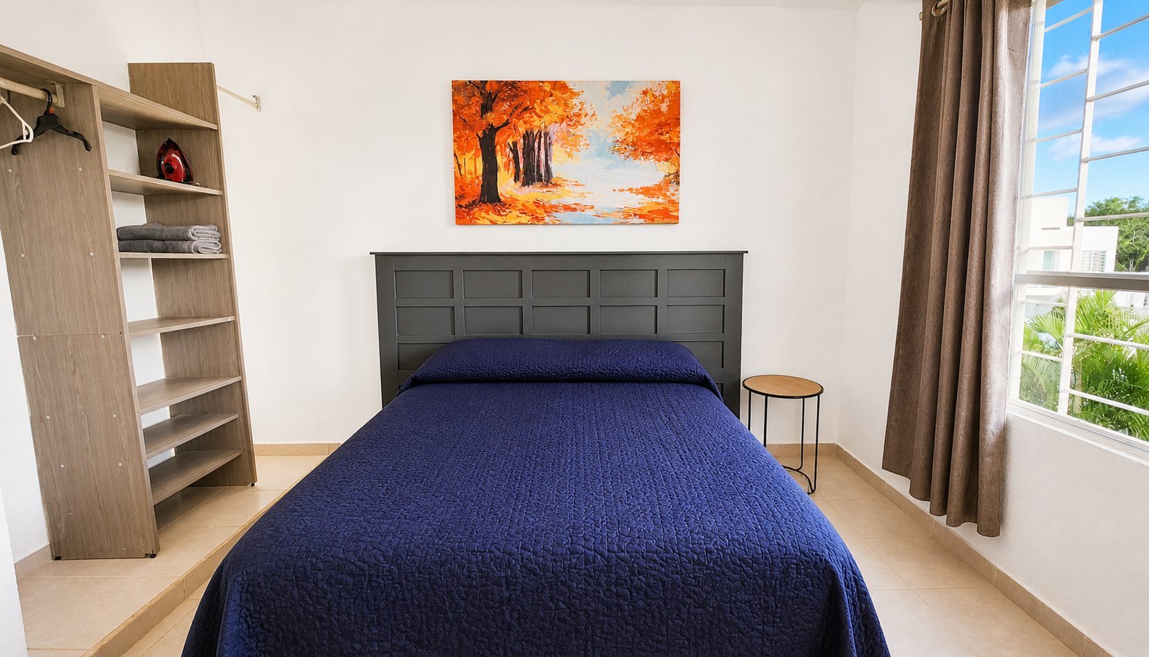 Modern bedroom with dark headboard, blue bedding, wood shelving, fall-themed artwork above bed, and large windows with beige curtains.