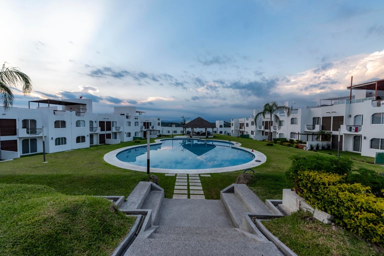 Modern white residential complex with curved blue swimming pool, lush green lawn, palm trees, and mountain backdrop at dusk