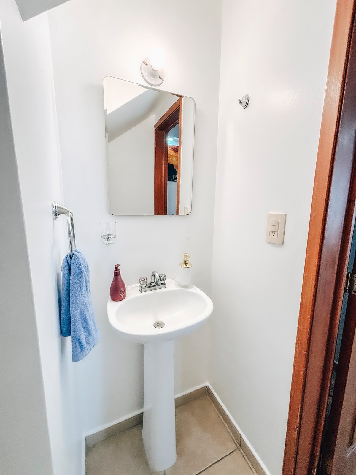Small bathroom with white pedestal sink, mirror, blue towel, and wooden door