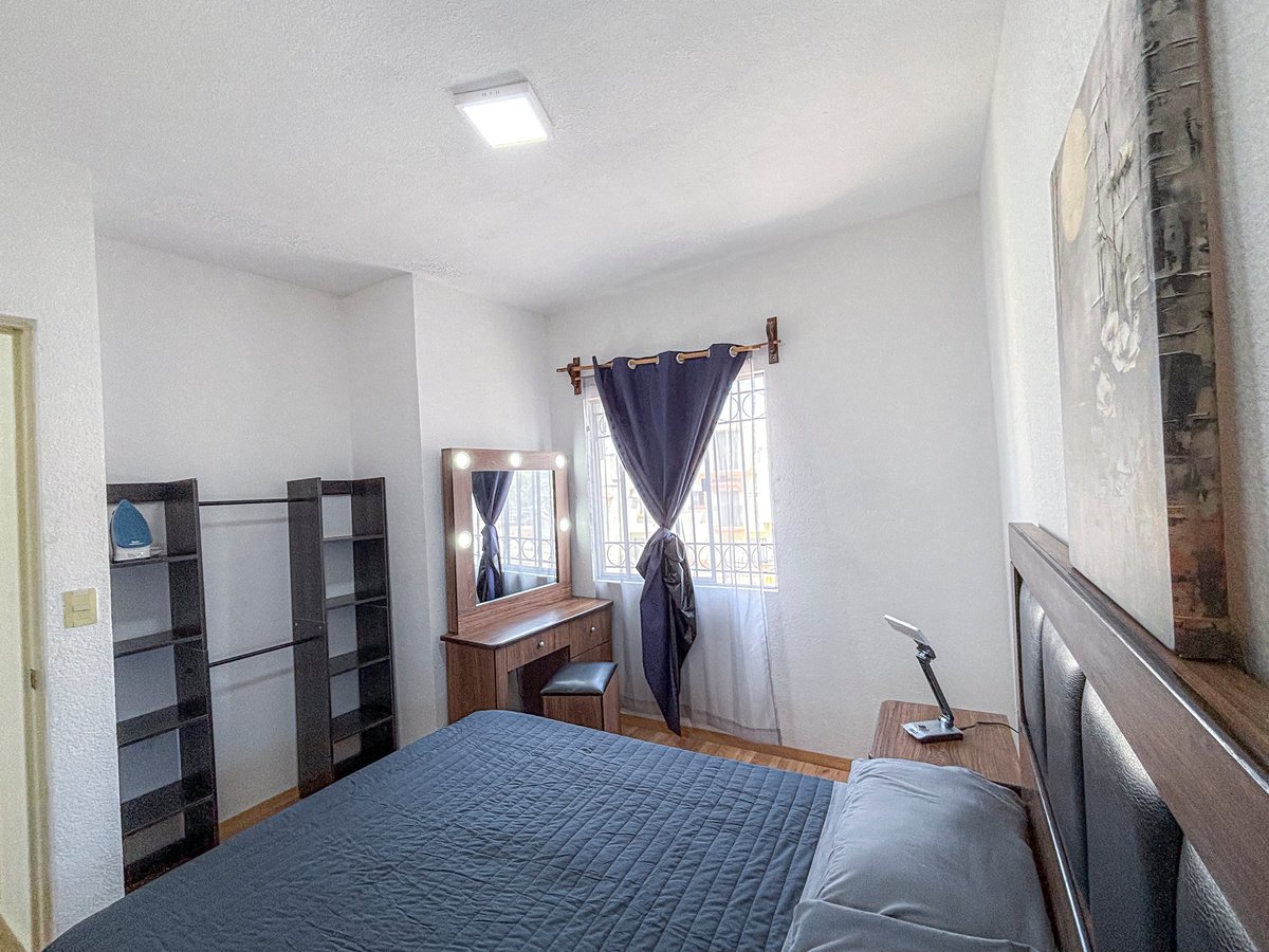 Bedroom interior with blue quilted bed, black shelving unit, wooden desk, and window with dark curtains