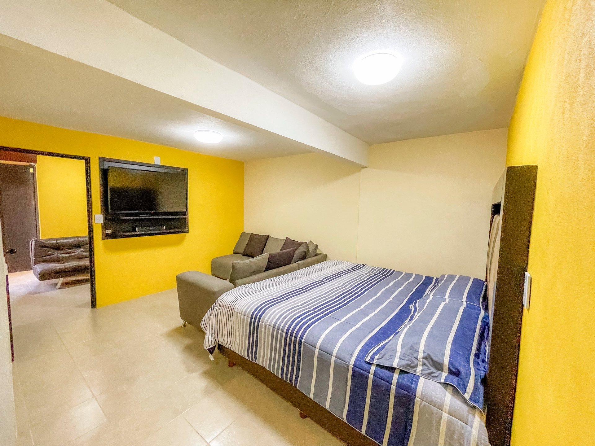 Modern bedroom with yellow walls, bed with blue striped bedding, gray couch, wall-mounted TV, and open doorway to adjoining room
