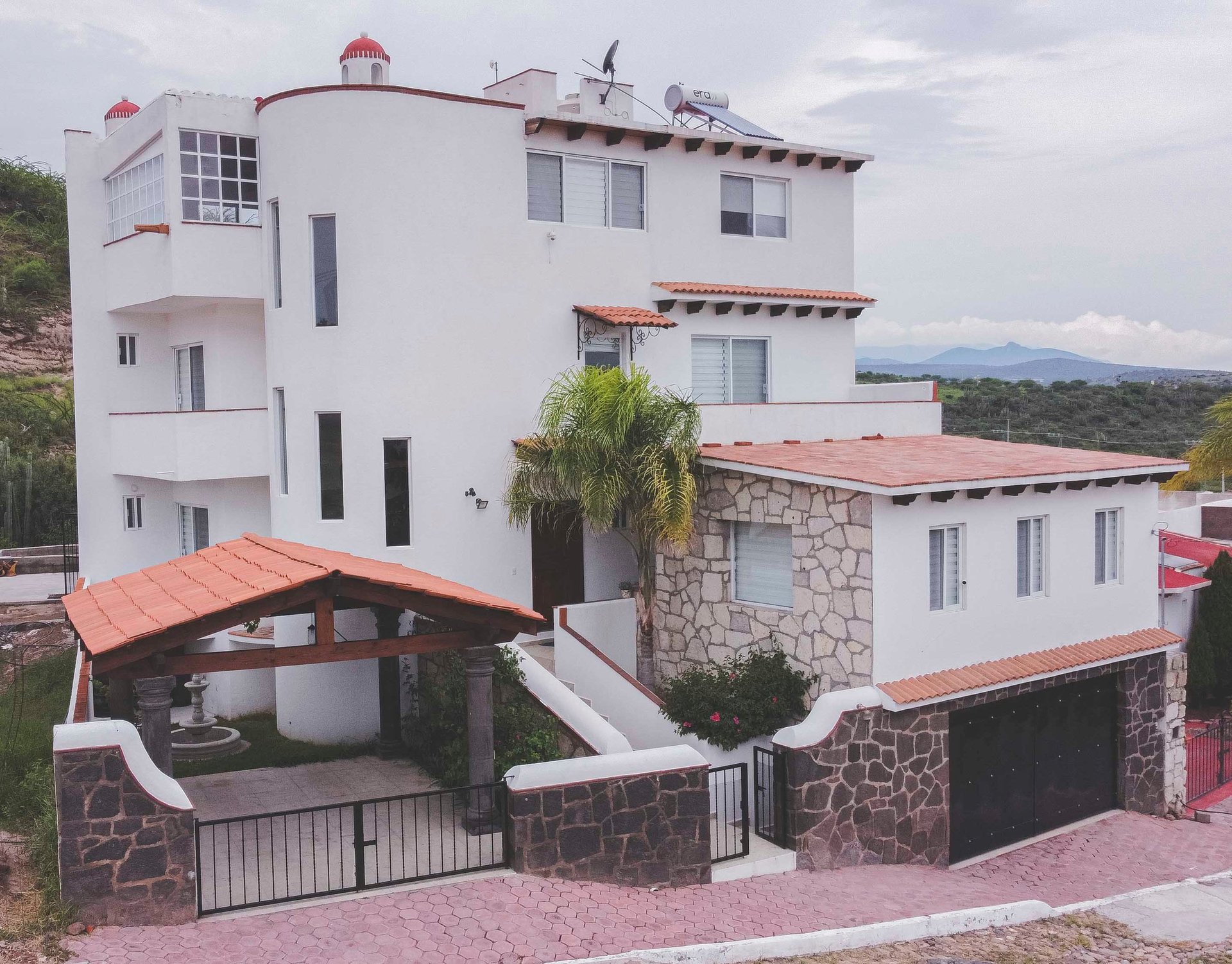 Modern white and stone hillside house with orange-tiled roofs, carport, palm tree, and mountain views in background