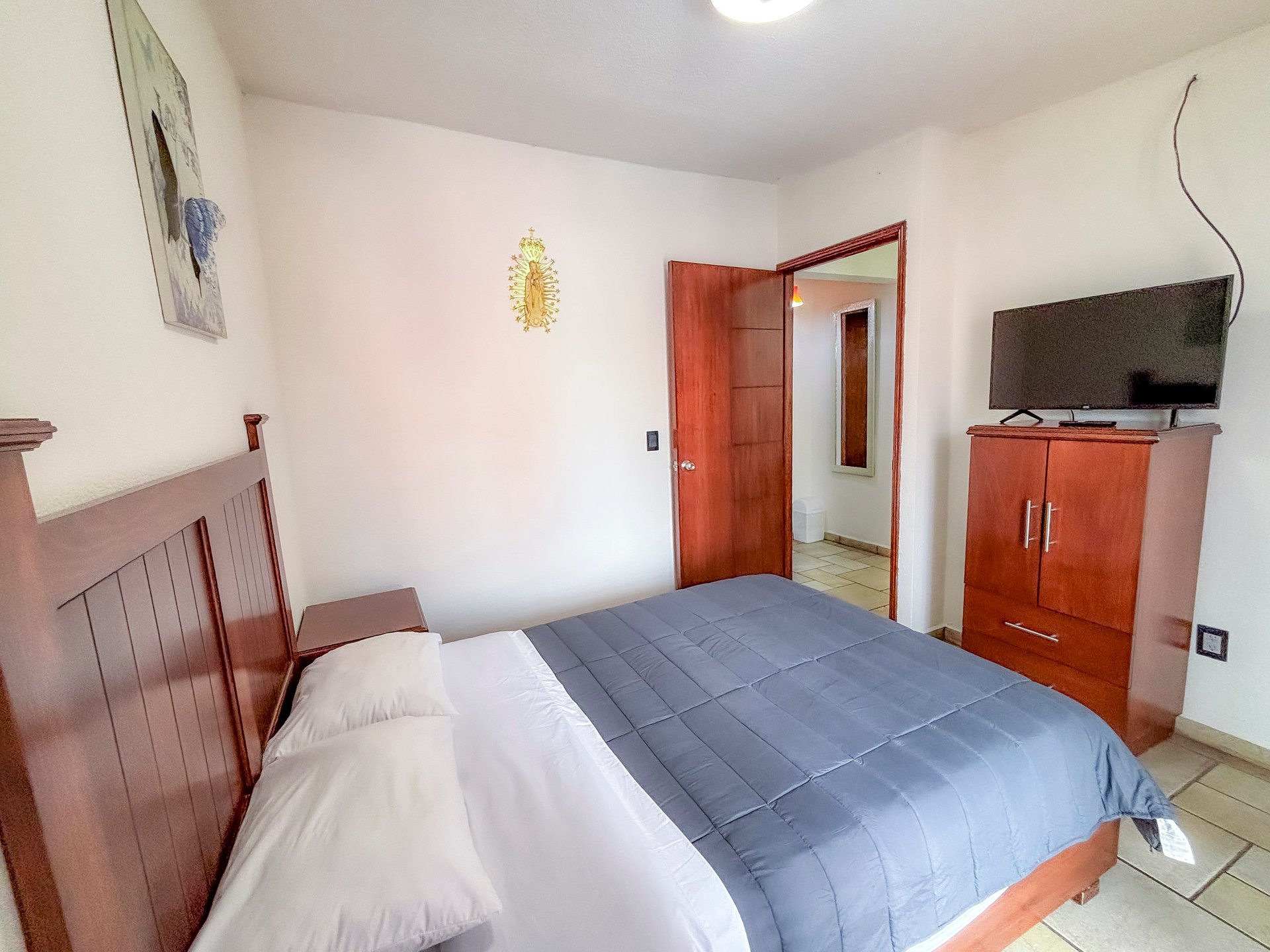 Modern bedroom with queen bed, wooden furniture, red wooden door, flat-screen TV mounted on dresser, and white walls with warm lighting.